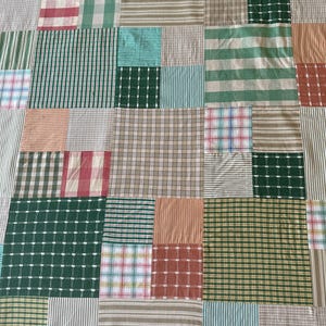 Op de afbeelding: Een patchwork quilt met een verscheidenheid aan stofpatronen en kleuren. De quilt bestaat uit rechthoekige blokken in tinten groen, roze, beige en blauw. Patronen zijn onder meer strepen, ruiten en effen kleuren, wat een interessant ontwerp creëert.