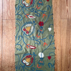 May include: Olive green fabric with embroidered floral patterns. The design features blue, red, yellow, and white floral and leaf motifs. The fabric is rectangular and appears to be a textile for home decor or clothing.