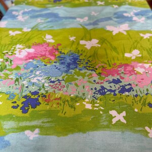 Könnte beinhalten: Nahaufnahme eines Stoffes mit Blumenmuster. Das Design zeigt ein leuchtend grünes Feld mit rosa, blauen und weißen Blumen und weißen Schmetterlingen. Hellblaue Bänder mit Wolken und weiteren Schmetterlingen trennen die Felder.