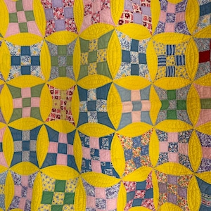 May include: A patchwork quilt featuring a repeating pattern of square blocks in various floral and geometric prints, separated by bright yellow curved fabric segments. The quilt displays a grid of multi-colored squares in shades of blue, pink, green, and purple, all stitched together with visible quilting lines.