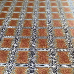 Könnte beinhalten: Nahaufnahme eines Stoffes mit Muster. Das Design zeigt ein Gitter aus rostfarbenen Quadraten, getrennt durch vertikale Blumenstreifen und horizontale weiß-schwarze Schachbrettlinien. Die floralen Elemente sind weiß.