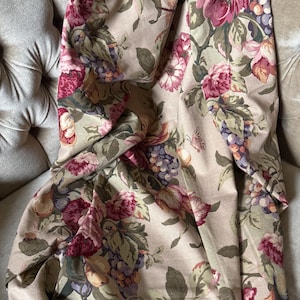 May include: A fabric drape featuring a beige background with a repeating pattern of deep red and pink roses, green leaves, and clusters of purple grapes. The textile has a soft, folded texture and displays a dense, colorful botanical print throughout.