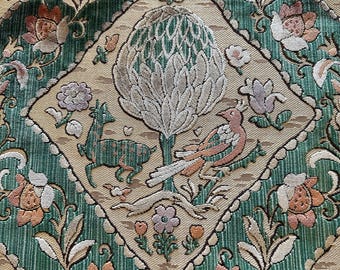 Può includere: Primo piano di un tessuto decorativo con un motivo ripetuto. Il design presenta una forma a diamante centrale con un albero, un uccello e un cervo. Lo sfondo è verde con motivi floreali e di foglie nei toni del pesca, crema e verde.