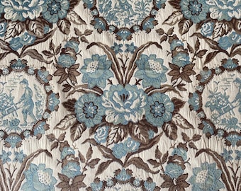 May include: Close-up of a floral fabric with a repeating pattern. The design features large, stylized blue flowers and brown leaves against a cream-colored background. Circular medallions with figures are interspersed throughout the pattern.