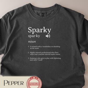 以下が含まれることがあります： 「Sparky」の文字と定義が白でプリントされた濃いグレーのTシャツ。定義は魔法使い、プロ、雷を扱う人について説明しています。Tシャツはクルーネックです。
