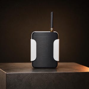 May include: A black and white electronic device with a black antenna. The device has a rectangular shape with rounded corners and white side brackets. It is set on a dark gray surface against a dark brown background.