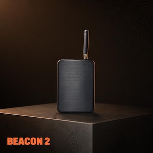 May include: A black rectangular device with a small antenna extending upwards. The device has a textured surface and is labeled "BEACON 2" in orange text at the bottom. The device is placed on a dark, square surface.