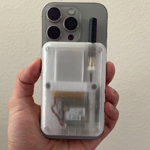 May include: A smartphone with a clear, rectangular case attached. The case has a small antenna and internal components visible through the transparent plastic. The phone's camera lenses are visible at the top.
