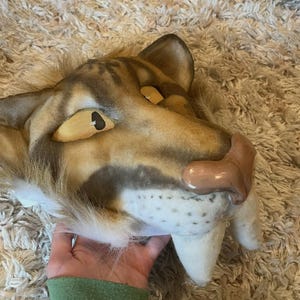 Könnte beinhalten: Eine detaillierte Maske eines Säbelzahntigers mit den Farben Beige, Braun und Weiß. Die Maske hat große, gebogene Fangzähne, eine rosa Nase und gelbe Augen mit schwarzen Pupillen. Die Maske besteht aus Stoff und Fell.