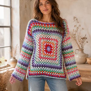 May include: A long-sleeved sweater featuring a multi-colored granny square crochet pattern. The garment displays a vibrant array of hues including red, orange, yellow, green, blue, and purple arranged in concentric squares, paired with light blue jeans.