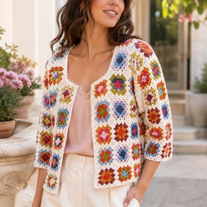Puede incluir: Una chaqueta corta de hilo color blanco roto con un patrón de cuadrados de ganchillo multicolor. La prenda tiene mangas tres cuartos, cuello redondo y diseño abierto en la parte delantera, sobre una camiseta rosa pálido y pantalones blancos.
