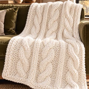 May include: A chunky, off-white cable knit throw blanket featuring thick, vertical braided patterns and a textured, heavy-gauge yarn construction. The blanket is draped over a surface, showcasing its intricate, raised knit design and soft, cream-colored appearance.