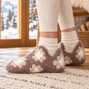 Puede incluir: Un par de zapatillas de ganchillo con un patrón de cuadrados de la abuela en marrón, beige y blanco. Las zapatillas se usan sobre calcetines acanalados de color crema, cubriendo los pies y tobillos con un diseño de punto texturizado y artesanal.
