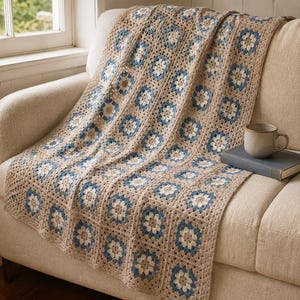 Peut inclure: Une couverture au crochet présentant un motif répétitif de carrés granny dans des tons beige, crème et bleu poussiéreux. La couverture est drapée sur un canapé de couleur neutre, avec un livre à couverture sombre et une tasse en céramique.