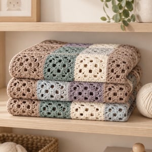 Può includere: Una coperta all'uncinetto piegata con un motivo ripetitivo a piastrelle granny in una palette di colori tenui composta da beige, verde salvia, crema e lavanda polveroso. La coperta è impilata in tre strati, mostrando la trama del filato.
