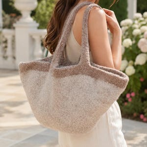 May include: A large, soft-textured tote bag featuring a two-tone design with a darker brown upper section and a lighter beige lower body. The bag has two integrated shoulder straps and a wide, open-top construction made of a fuzzy, wool-like material.