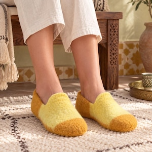 Puede incluir: Un par de zapatillas tipo pantufla con un diseño de dos tonos, con una sección central en amarillo brillante y una puntera y talón en amarillo mostaza más oscuro. Las zapatillas tienen una textura suave y afieltrada con punta redondeada.