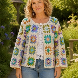 May include: A cream-coloured crochet jacket featuring colourful square patterns in shades of blue, green, orange, and purple. The jacket has long sleeves and a button closure. The person is wearing a white top and blue jeans.
