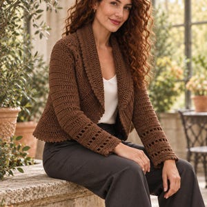 May include: A brown crochet cardigan featuring a wide shawl collar, long sleeves with a decorative openwork stitch pattern at the cuffs, and a scalloped hem. The garment is worn over a white camisole and dark grey trousers.