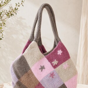May include: A soft, fuzzy tote bag featuring a diamond-patterned patchwork design in shades of magenta, light pink, lavender, and brown. The bag has two integrated handles and is decorated with small, embroidered flower motifs on the panels.