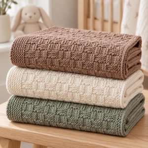 Könnte beinhalten: Ein Stapel aus drei gefalteten Strickdecken in den Farben Braun, Creme und Salbeigrün. Jede Decke weist ein durchgehendes Korbgeflecht-Strukturmuster im Gewebe auf, mit sauberen Kanten und einem gleichmäßigen Erscheinungsbild des Strickmusters.