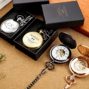 Pode incluir: Relógios de bolso em caixas pretas. Um relógio é prateado com um design de chifre de veado e as iniciais "CK". Outro relógio é dourado com o nome "ADRIAN" e o texto "Groomsman 12/05/25". Dois relógios de bolso abertos mostram o mecanismo interno, um preto e outro dourado.