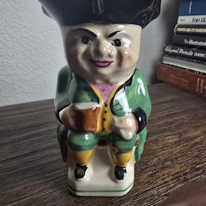 Könnte beinhalten: Keramikfigur eines Mannes mit Dreispitz und grünem Mantel, der auf einer Bank sitzt. Die Figur hat ein lächelndes Gesicht und hält einen Krug. Der Sockel ist cremefarben, die Figur ist etwa 15 cm hoch. Bücher im Hintergrund.