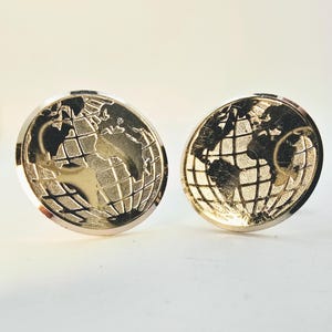 Puede incluir: Un par de gemelos dorados con un diseño detallado de globo terráqueo. Los gemelos tienen forma circular con un patrón de mapa del mundo en relieve y un borde plateado. El diseño es intrincado y el aspecto general es elegante.