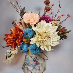 May include: A decorative floral arrangement in a vintage-style metal container. The bouquet features orange, peach, and cream-colored flowers, with blue pumpkin accents. The container has a floral and cherub design.