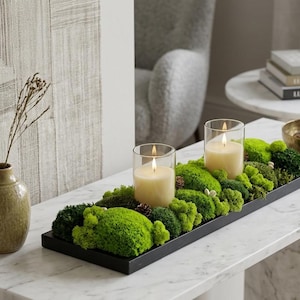 May include: A decorative centerpiece featuring two lit candles in glass holders, set on a bed of green moss and small pinecones within a black rectangular tray. A vase with a dried plant is on the left.