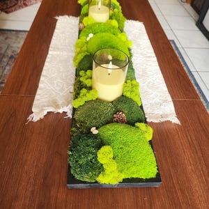 May include: A rectangular, black wooden centerpiece with vibrant green moss and two glass candle holders. The candles are lit, and the arrangement includes small pinecones and decorative elements, creating a natural, earthy aesthetic.