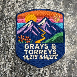 May include: An embroidered patch featuring a mountain landscape with a yellow sun, purple peaks, a winding orange trail, and orange flowers. The text reads GRAYS & TORREYS 14,275' & 14,272' in white lettering against a dark blue background.