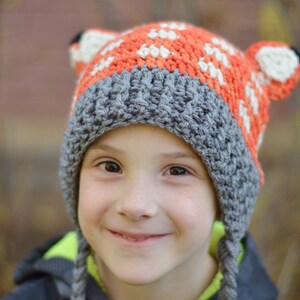 May include: A child wearing a grey and orange crocheted hat with fox ears. The hat has a white stripe and is pulled down over the child's forehead. The child is smiling and looking at the camera.
