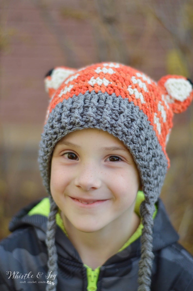 May include: A child wearing a grey and orange crocheted hat with fox ears. The hat has a white stripe and is pulled down over the child's forehead. The child is smiling and looking at the camera.