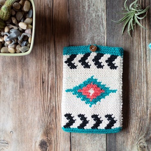 Puede incluir: Una funda para teléfono de ganchillo con un patrón geométrico en tonos turquesa, blanco, negro y rojo. La funda tiene un cierre de botón.