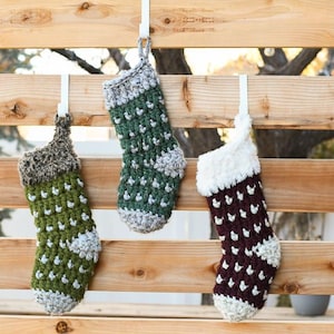 May include: Three crocheted Christmas stockings hanging on a wooden fence. The stockings are green, brown, and white with a white pom-pom on each.