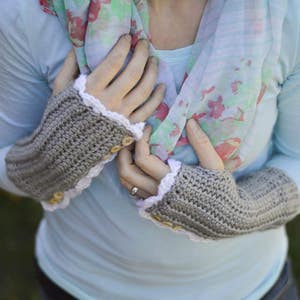 May include: A pair of gray crocheted fingerless gloves with white lace trim and wooden buttons. The gloves are worn by a person wearing a light blue shirt and a floral scarf.