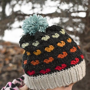May include: A black crochet hat with a colourful heart pattern and a light blue pompom on top. The hat has a grey brim.