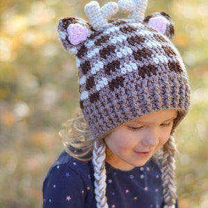 May include: A brown and white crocheted deer hat with pink ears and braided ties. The hat is worn by a child with blonde hair and a blue shirt with white stars.