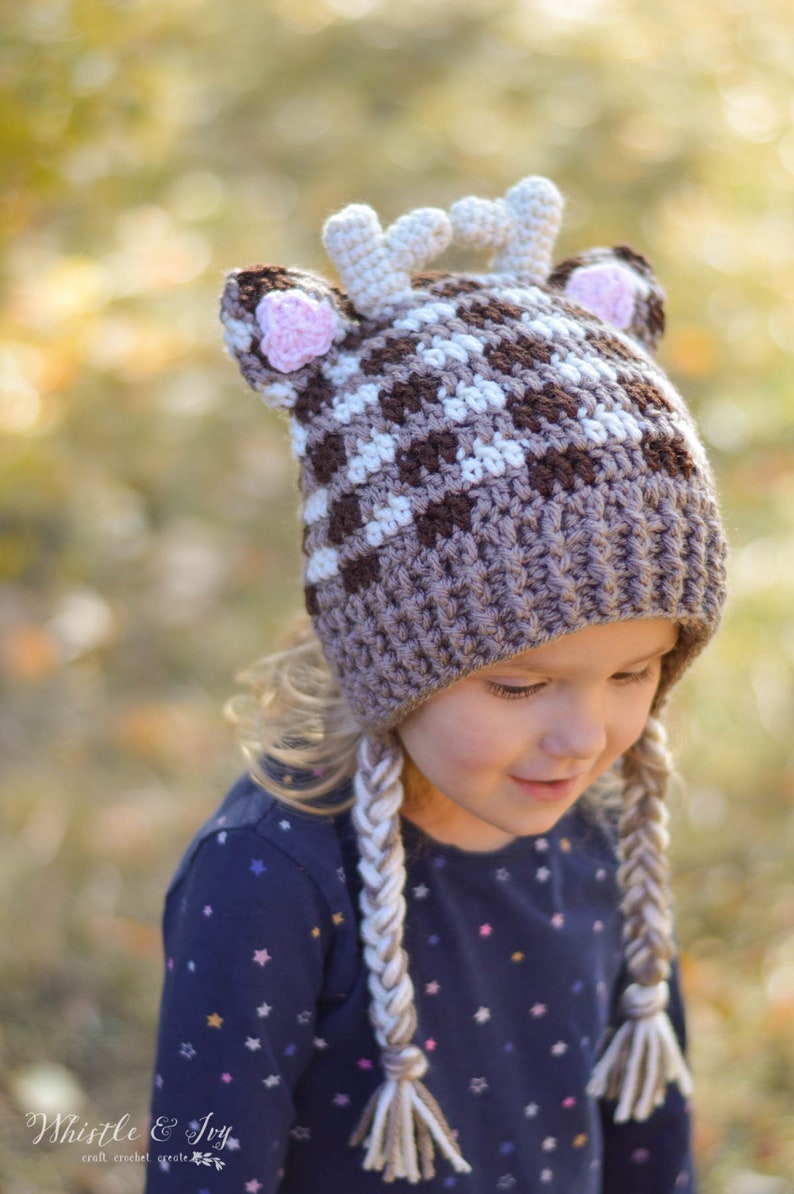 May include: A brown and white crocheted deer hat with pink ears and braided ties. The hat is worn by a child with blonde hair and a blue shirt with white stars.