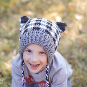 May include: A child wearing a grey and white crocheted hat with raccoon ears. The hat has a black and white striped trim. The child is smiling and looking at the camera.