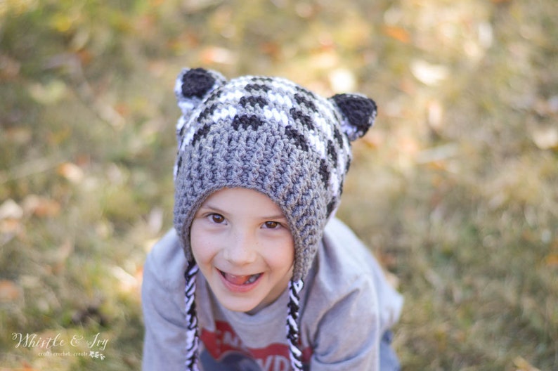 May include: A child wearing a grey and white crocheted hat with raccoon ears. The hat has a black and white striped trim. The child is smiling and looking at the camera.