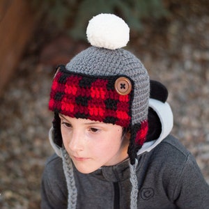 May include: A grey and red crocheted hat with ear flaps and a white pom pom on top. The hat has a black and red plaid pattern on the ear flaps and a brown button on the side.