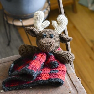 May include: A brown crocheted moose with white antlers is holding a red and black plaid crocheted blanket. The moose and blanket are sitting on a rusty metal chair.