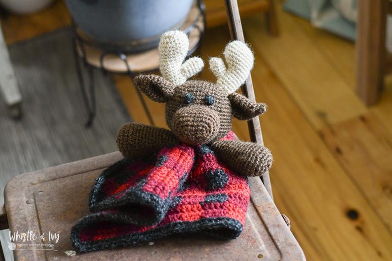 May include: A brown crocheted moose with white antlers is holding a red and black plaid crocheted blanket. The moose and blanket are sitting on a rusty metal chair.