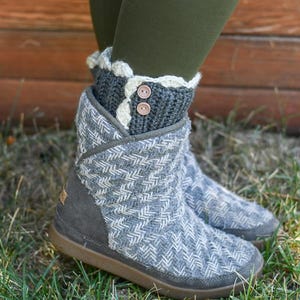 May include: Gray and white knitted ankle boots with a white crocheted cuff and two brown buttons. The boots are worn by a person standing on grass.