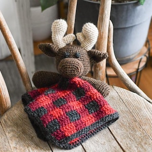 May include: A brown crocheted moose with beige antlers holding a red and black plaid blanket. The moose and blanket are sitting on a wooden chair.