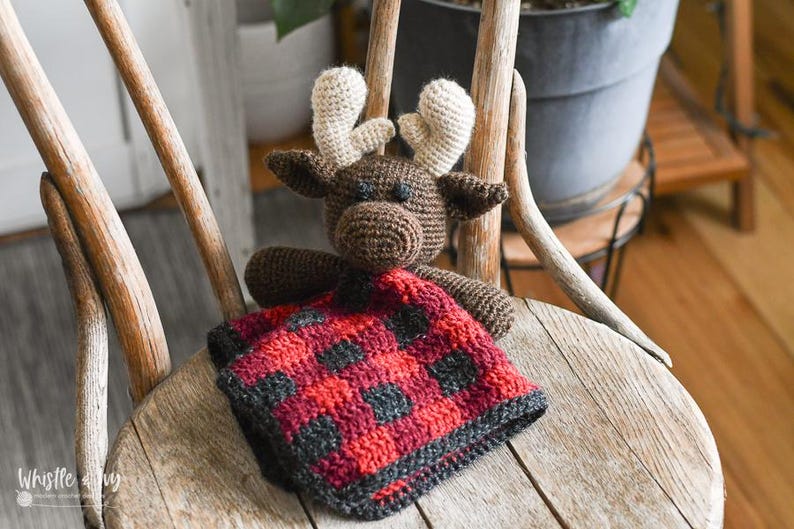 May include: A brown crocheted moose with beige antlers holding a red and black plaid blanket. The moose and blanket are sitting on a wooden chair.