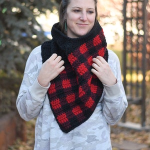 Op de afbeelding: Een zwarte en rode geruite gehaakte cowl-hals sjaal. De sjaal wordt gedragen door een persoon die voor een wazige achtergrond staat.