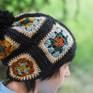 Könnte beinhalten: Eine schwarze Häkelmütze mit einem Bommel oben. Die Mütze hat ein geometrisches Muster mit Quadraten in verschiedenen Farben, darunter Braun, Orange, Blau und Grün.